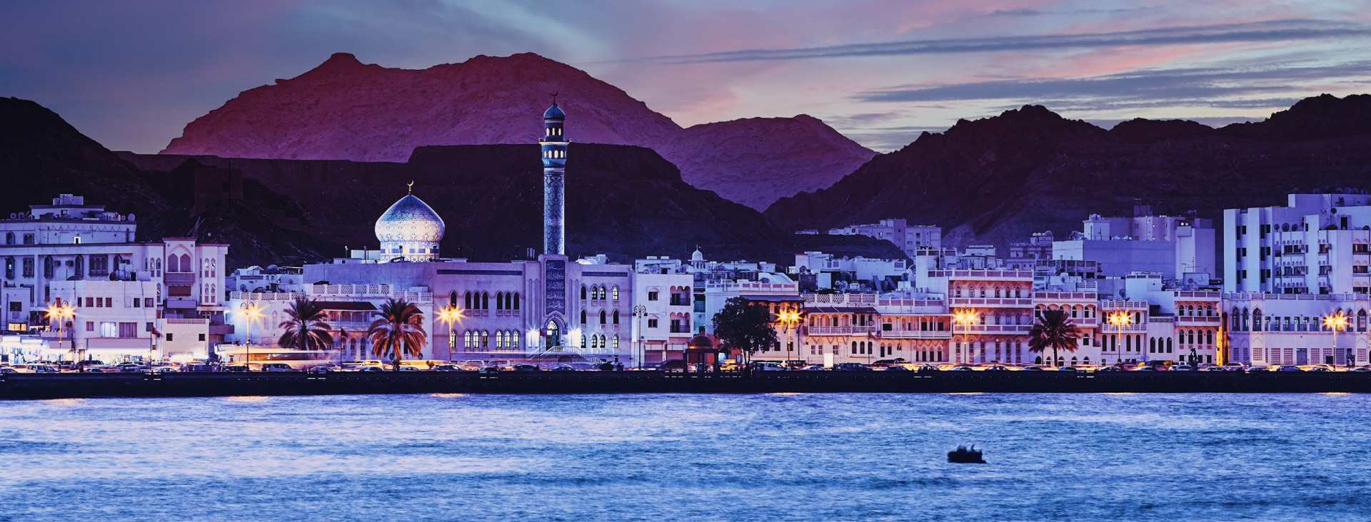 The Muscat waterfront at dusk, with a blue-domed mosque and minaret in front of the mountains
